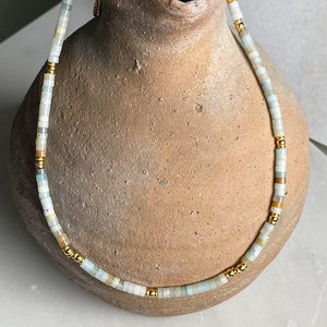 May include: A delicate necklace made of light blue and beige beads with gold accents. The necklace is displayed on a brown ceramic vase.