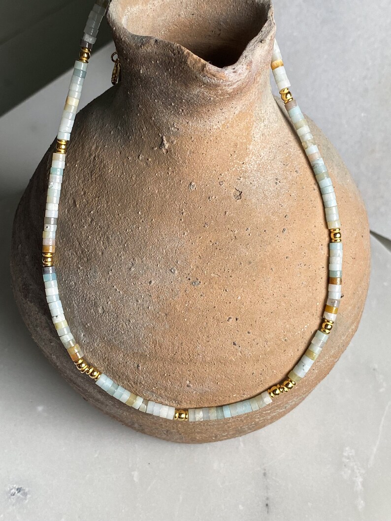 May include: A delicate necklace made of light blue and beige beads with gold accents. The necklace is displayed on a brown ceramic vase.