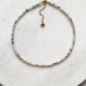 May include: A delicate gold chain necklace with small, light blue, white, and beige beads. The necklace has a small gold clasp.