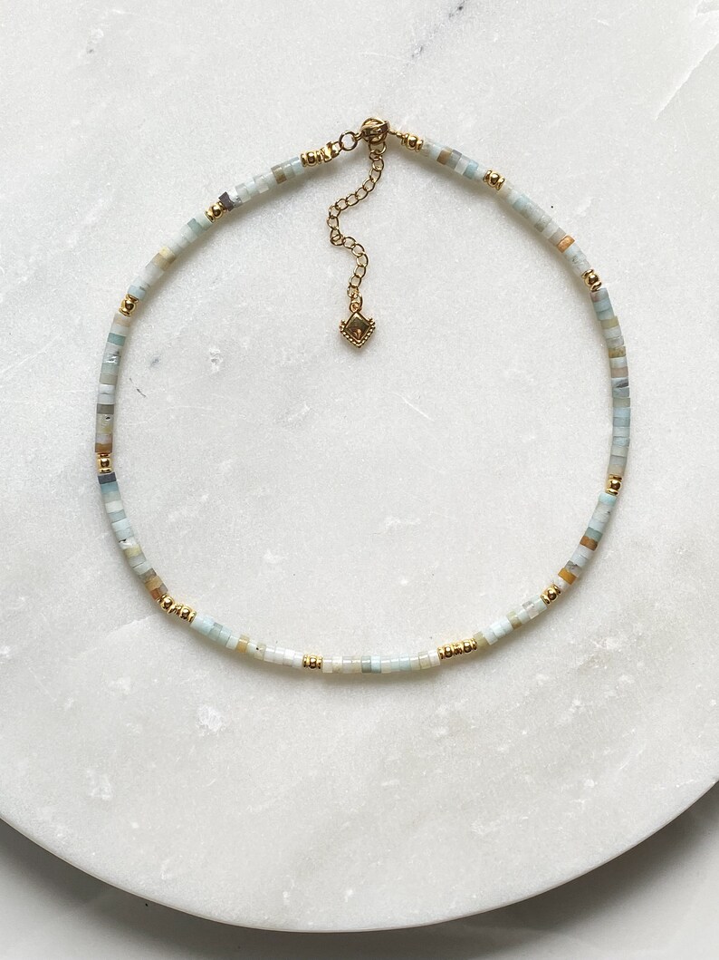May include: A delicate gold chain necklace with small, light blue, white, and beige beads. The necklace has a small gold clasp.