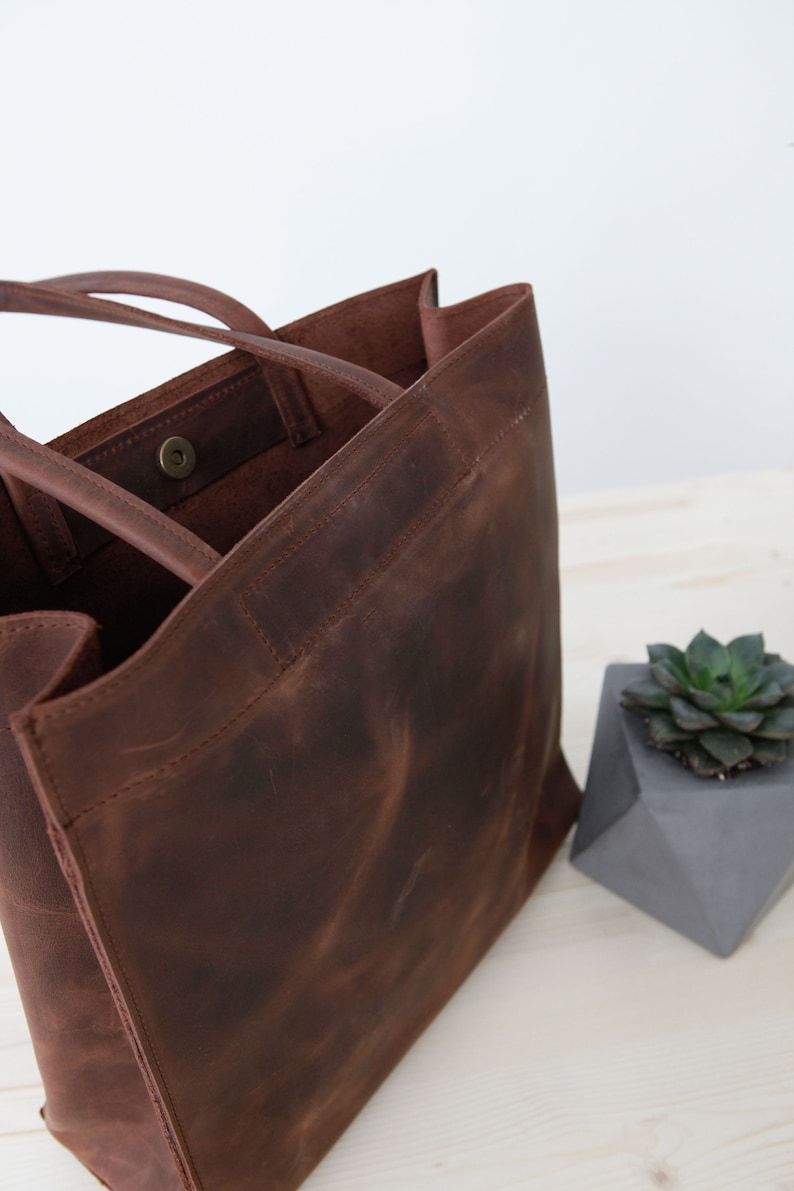 May include: A brown leather tote bag with a single snap closure. The bag has two handles and a large, open top.