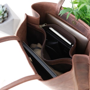 May include: A brown leather tote bag with a zippered pocket and a flap closure. The bag is open and has a laptop, a notebook, and other items inside.