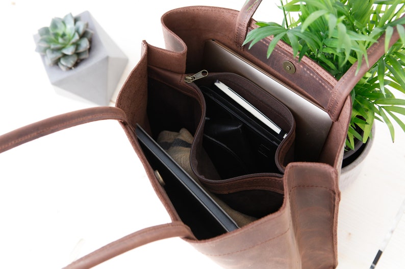 May include: A brown leather tote bag with a zippered pocket and a flap closure. The bag is open and has a laptop, a notebook, and other items inside.