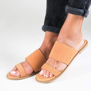 May include: A pair of tan leather sandals with two straps across the foot. The sandals have a flat sole and are worn by a person with bare feet.