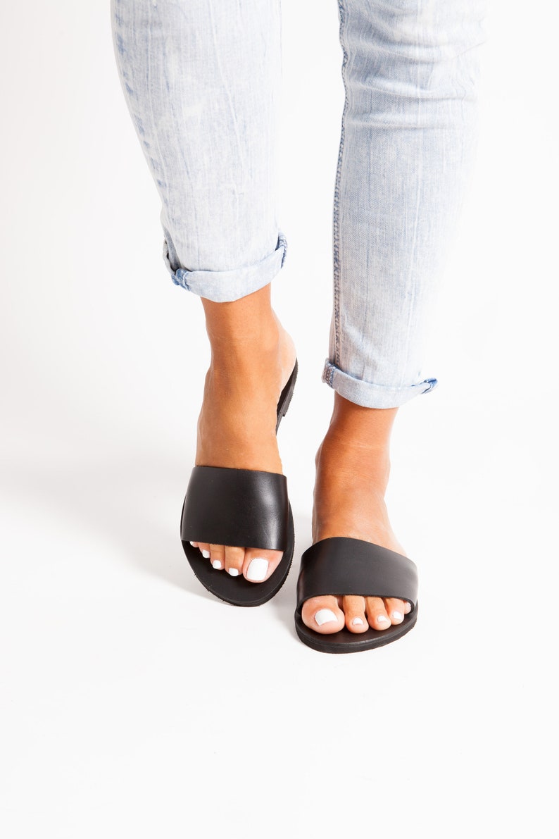 May include: Black leather slide sandals with a wide strap, worn with light blue denim jeans. The sandals have a simple, minimalist design. The jeans are cuffed at the ankles.