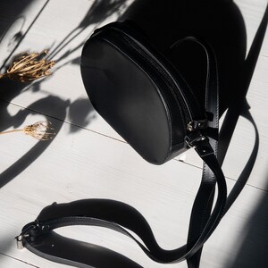 May include: A black leather crossbody bag with a round shape and a long adjustable strap. The bag is laying on a white wooden surface.