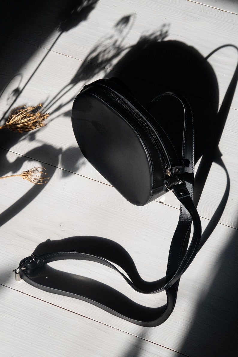 May include: A black leather crossbody bag with a round shape and a long adjustable strap. The bag is laying on a white wooden surface.