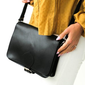 Puede incluir: Un bolso de cuero negro con cierre de hebilla plateada. El bolso se lleva al hombro y tiene una correa.