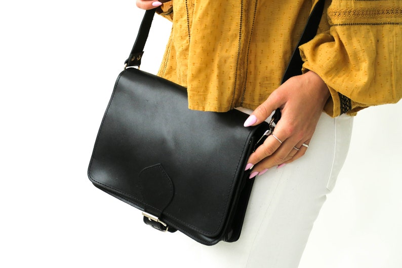 Puede incluir: Un bolso de cuero negro con cierre de hebilla plateada. El bolso se lleva al hombro y tiene una correa.