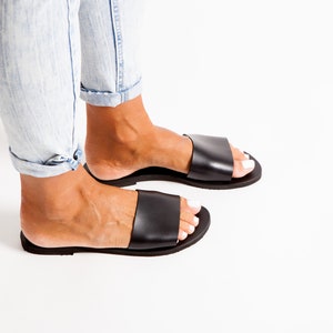 May include: Black leather slide sandals. The sandals have a wide strap across the top of the foot and a flat sole. The person is wearing light blue jeans. The sandals are a casual footwear option.
