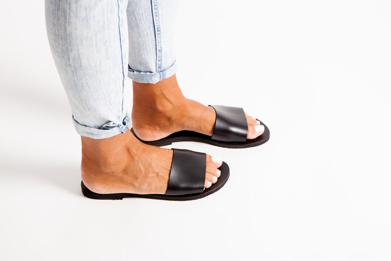 May include: Black leather slide sandals. The sandals have a wide strap across the top of the foot and a flat sole. The person is wearing light blue jeans. The sandals are a casual footwear option.