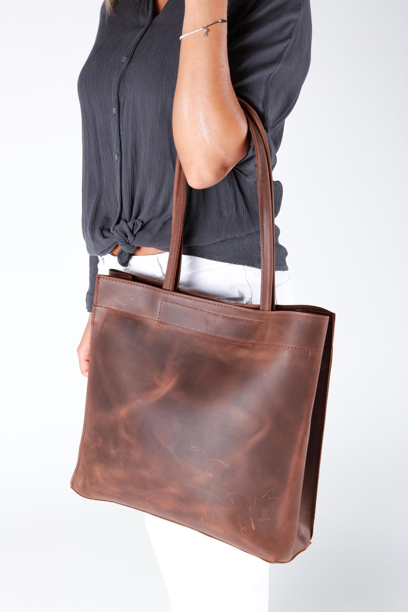 May include: A brown leather tote bag with a distressed finish. The bag has a wide, flat base and two long handles.
