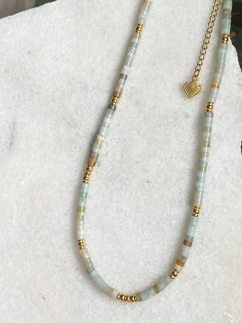 May include: A delicate gold chain necklace with a heart charm and a strand of light blue, white, and brown beads.