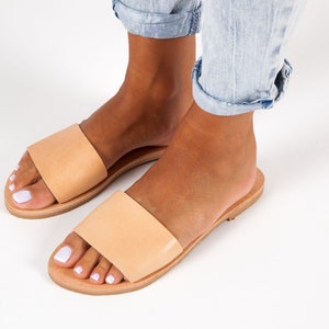 May include: A pair of light tan leather slide sandals. The sandals have a wide strap across the top of the foot and a flat sole. The sandals are shown on a white background.