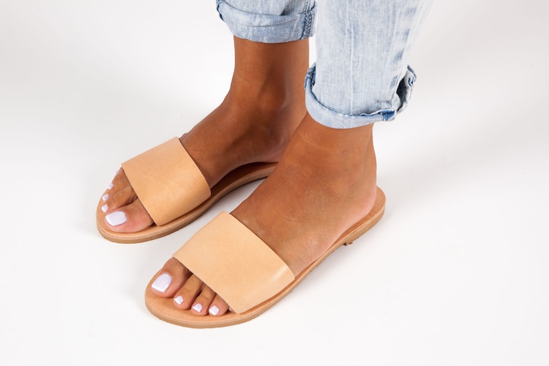 May include: A pair of light tan leather slide sandals. The sandals have a wide strap across the top of the foot and a flat sole. The sandals are shown on a white background.