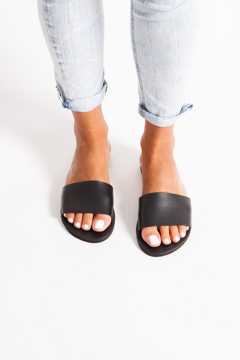 May include: Black leather slide sandals with a wide strap, worn with light blue denim jeans. The sandals are on a white background. The jeans are cuffed at the ankles.