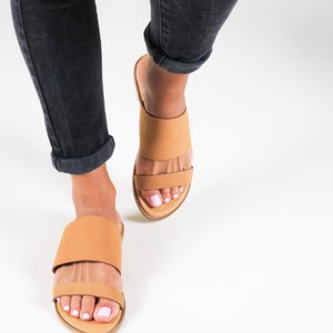 May include: A pair of tan leather slide sandals with two straps across the foot. The sandals are worn by a person wearing dark denim jeans.