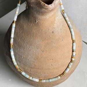 May include: A delicate necklace made of light blue, white, and beige beads with gold accents. The necklace is displayed on a brown ceramic vase.