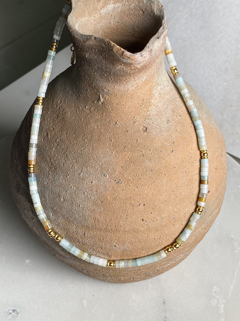 May include: A delicate necklace made of light blue, white, and beige beads with gold accents. The necklace is displayed on a brown ceramic vase.