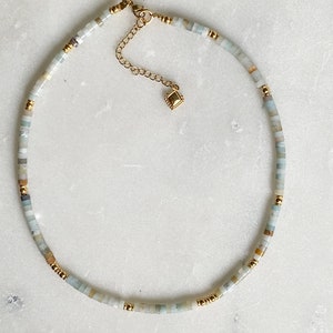 May include: A delicate gold chain necklace with a small square charm. The necklace is made with light blue, white, and brown beads.
