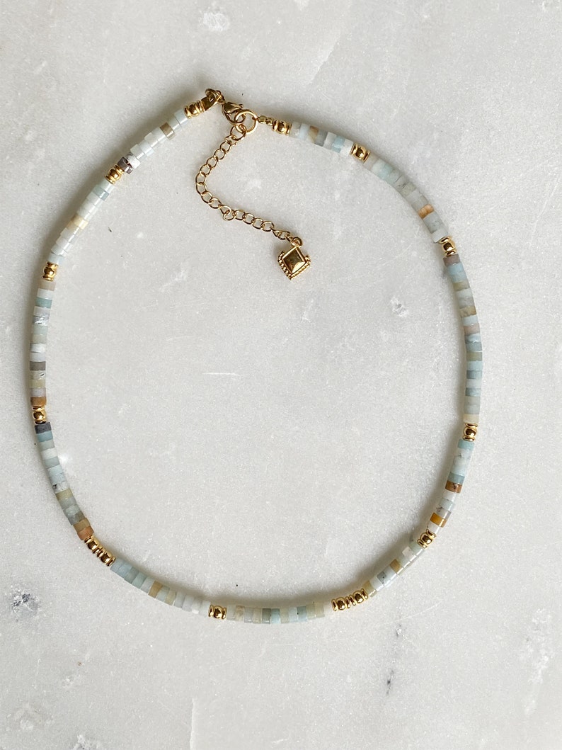 May include: A delicate gold chain necklace with a small square charm. The necklace is made with light blue, white, and brown beads.