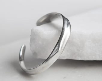 May include: A silver-toned cuff bracelet with an open design and a smooth, slightly irregular, organic surface texture. The metal band features a polished finish that reflects light, creating subtle highlights along its curved, rounded form.