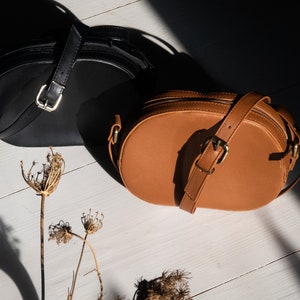 May include: Two round leather handbags, one black and one brown, on a white wooden surface. Dried flowers are in the foreground.