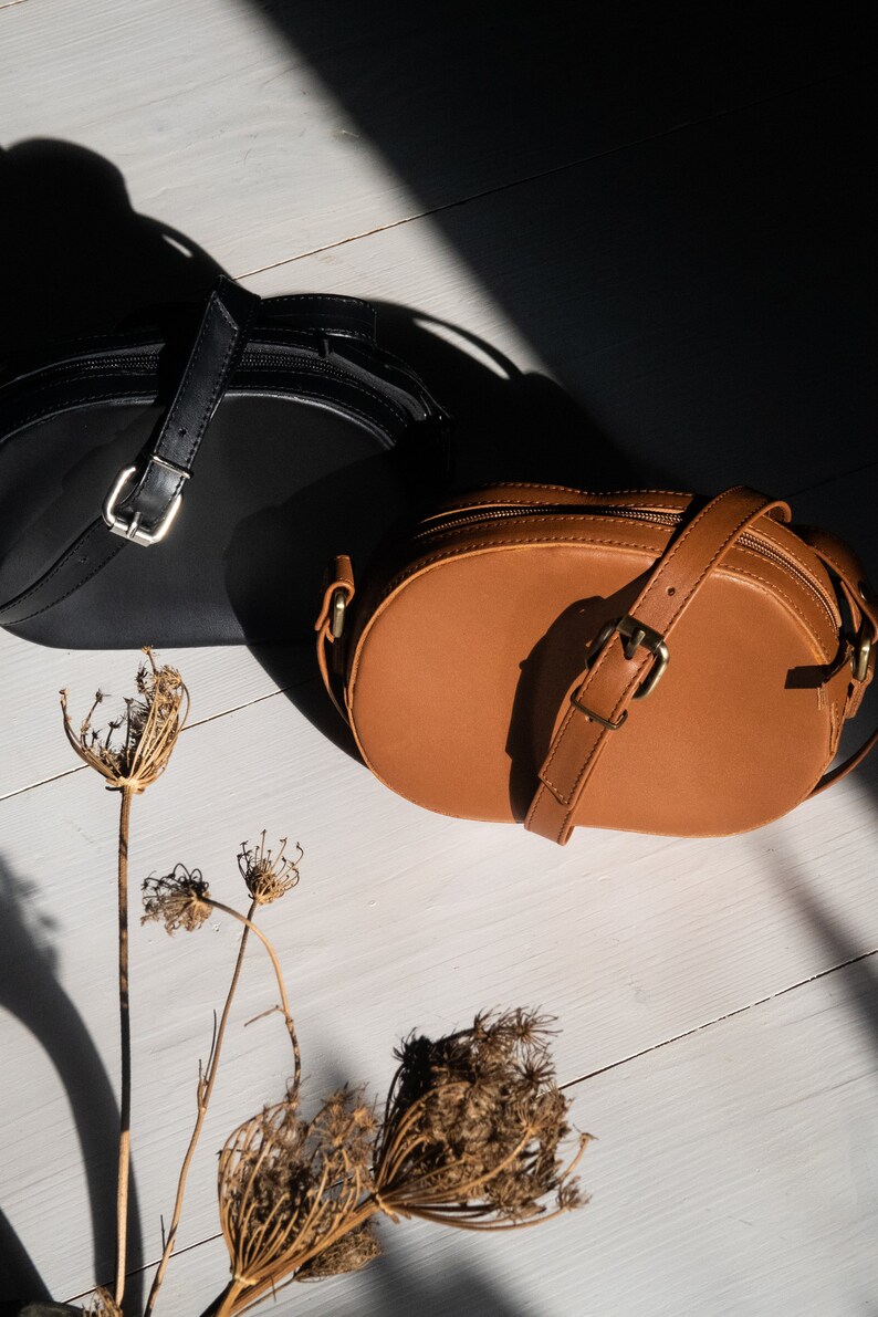 May include: Two round leather handbags, one black and one brown, on a white wooden surface. Dried flowers are in the foreground.