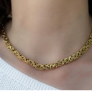 May include: A gold-coloured necklace featuring a Byzantine chain design. The necklace is made of interlocking links and is worn around the neck. The necklace is a jewellery item.