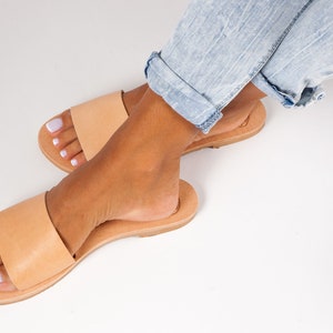 May include: A pair of light brown leather sandals with a single strap across the top. The sandals are on a light brown sole. The image shows the sandals on a white surface with a person wearing light blue jeans.