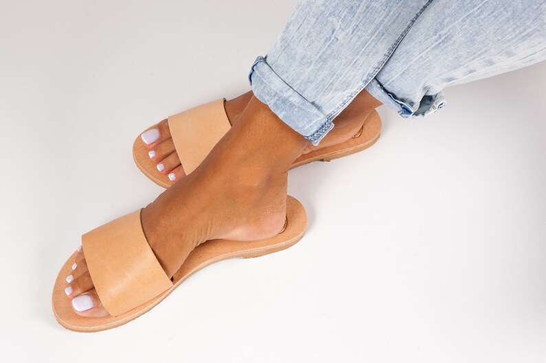 May include: A pair of light brown leather sandals with a single strap across the top. The sandals are on a light brown sole. The image shows the sandals on a white surface with a person wearing light blue jeans.