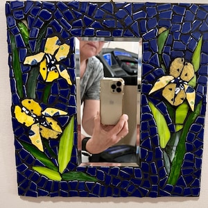 May include: Square mosaic mirror with a blue tile border. Yellow and white flower designs are featured on the corners. Green leaves are interspersed throughout the design. The mirror is rectangular.