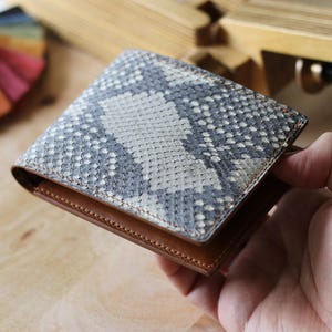 Peut inclure: Un portefeuille à deux volets avec un motif peau de serpent dans les tons de gris et de blanc. Le portefeuille a un intérieur et une garniture extérieure en cuir marron. Le portefeuille est tenu dans une main.
