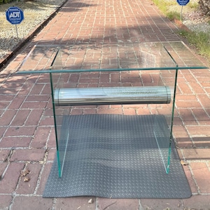 May include: A glass table with a chrome metal support bar. The table has a clear glass top and a clear glass base. The table is sitting on a black mat.