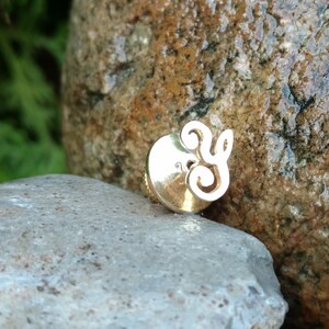May include: A gold-colored metal pin with a decorative swirl design. The pin is sitting on a gray rock.