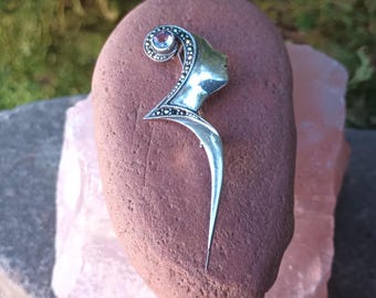 May include: Silver-toned brooch featuring a profile of a woman's face with a swirling design and a single gemstone. The brooch has a long, pointed tail and is set on a reddish-brown stone.