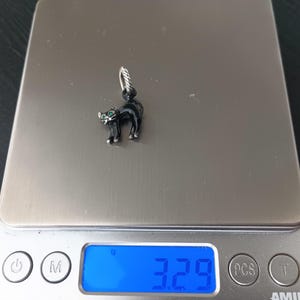 May include: A black cat charm with green eyes and silver accents. The cat is standing on a digital scale, which reads 3.29 grams. The charm has a silver loop for attaching to a bracelet or necklace.