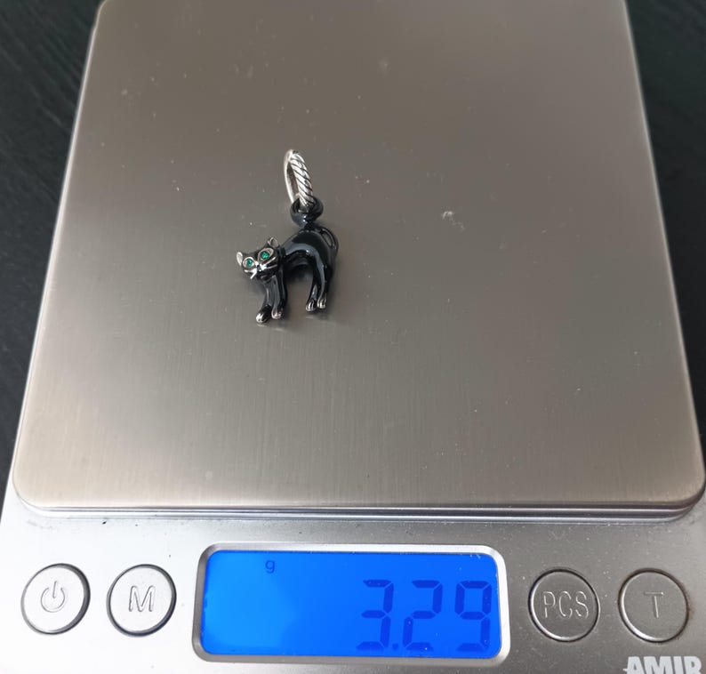 May include: A black cat charm with green eyes and silver accents. The cat is standing on a digital scale, which reads 3.29 grams. The charm has a silver loop for attaching to a bracelet or necklace.