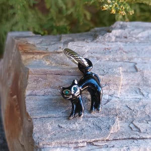 May include: A black cat charm with green eyes and a silver-colored ring. The cat is standing with its back arched. The charm is made of a shiny, reflective material.