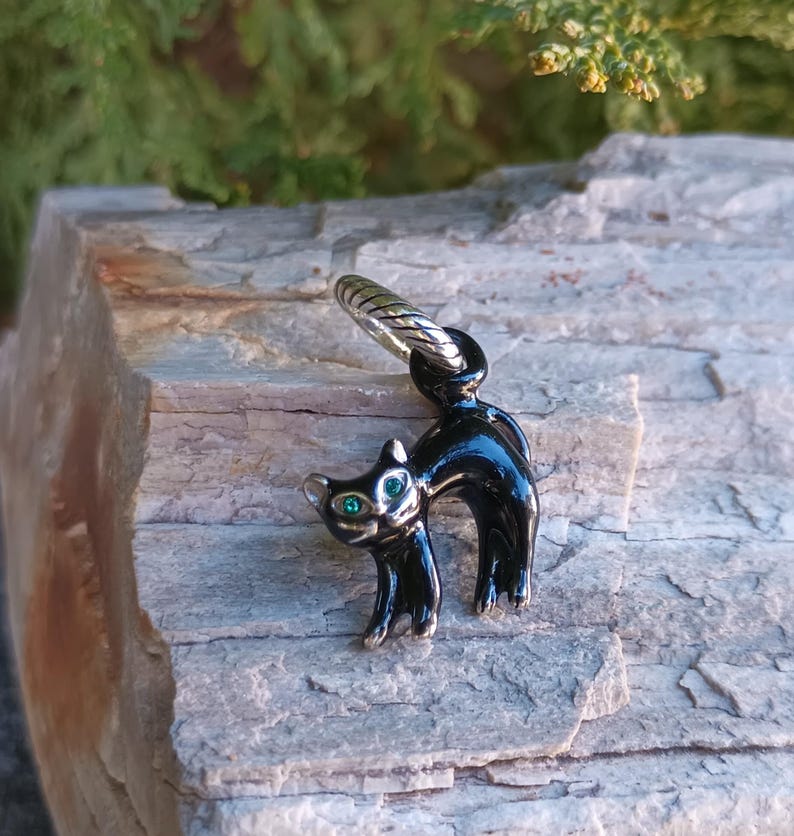 May include: A black cat charm with green eyes and a silver-colored ring. The cat is standing with its back arched. The charm is made of a shiny, reflective material.