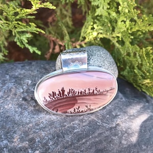 May include: Oval pendant with a silver setting. The pendant features a landscape design in shades of pink, brown, and white. The silver bail is decorated with an etched pattern. The pendant is displayed on a gray stone.