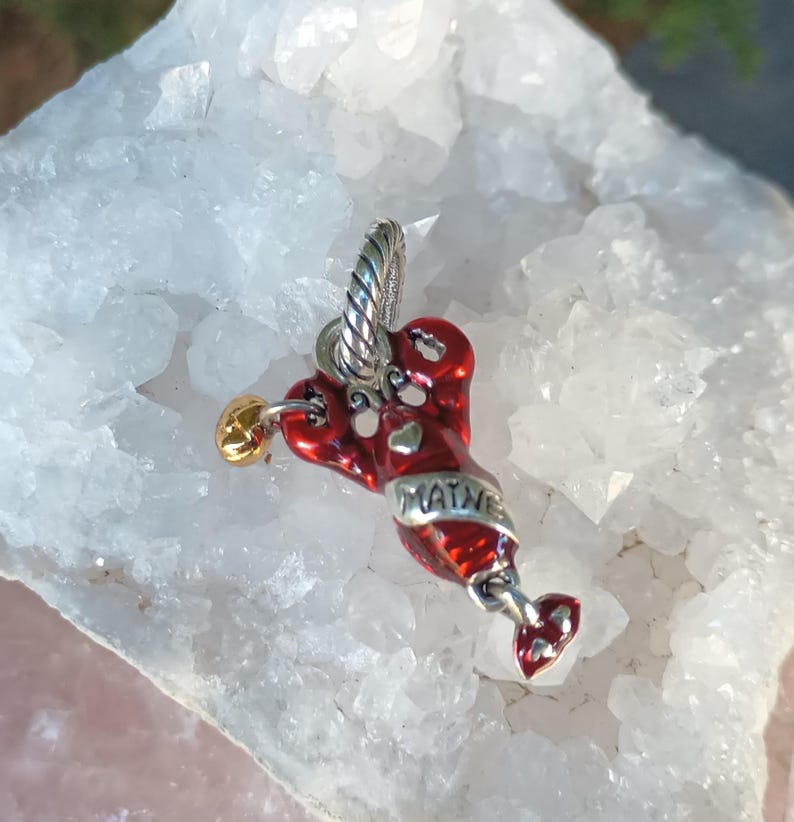K&ouml;nnte beinhalten: Ein roter Hummer-Anh&auml;nger mit dem Wort "MAINE" in silbernen Buchstaben. Der Hummer hat herzf&ouml;rmige Akzente und eine goldfarbene Kugel. Der Anh&auml;nger h&auml;ngt an einer silberfarbenen &Ouml;se.