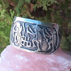 May include: A wide, silver-colored cuff bracelet with a detailed relief design. The relief depicts figures in a stylized, possibly historical, scene. The bracelet is displayed on a pink, rough-textured stone, with green foliage in the background.