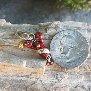 K&ouml;nnte beinhalten: Ein roter und silberner Hummer-Anh&auml;nger mit dem Wort "MAINE" darauf, neben einem US-Vierteldollar. Der Hummer-Anh&auml;nger hat einen goldfarbenen Hammer und eine silberne Schlaufe zum Befestigen an einem Armband oder einer Halskette. Der Anh&auml;nger liegt auf einem Holzst&uuml;ck.