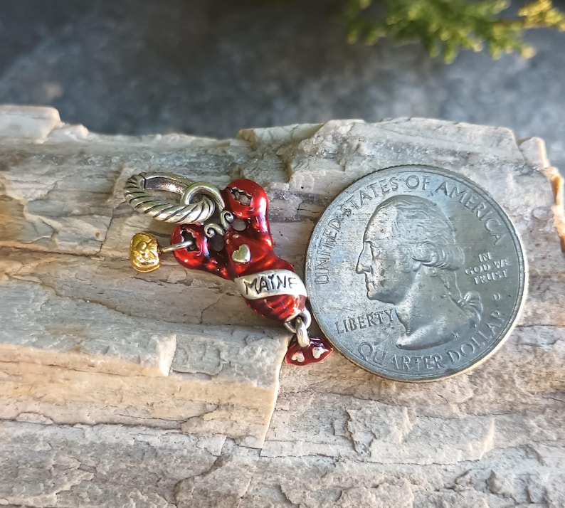K&ouml;nnte beinhalten: Ein roter und silberner Hummer-Anh&auml;nger mit dem Wort "MAINE" darauf, neben einem US-Vierteldollar. Der Hummer-Anh&auml;nger hat einen goldfarbenen Hammer und eine silberne Schlaufe zum Befestigen an einem Armband oder einer Halskette. Der Anh&auml;nger liegt auf einem Holzst&uuml;ck.