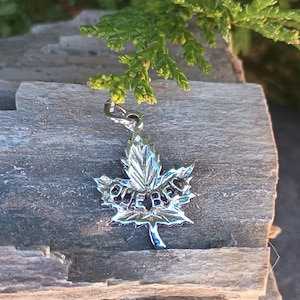 May include: A silver-colored maple leaf charm with the word "QUEBEC" engraved on it. The charm is suspended from a small loop and hangs against a weathered wooden surface. Green foliage is visible in the background.