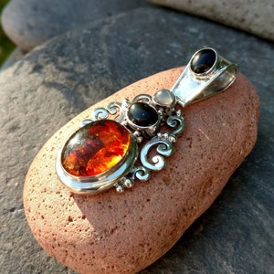 May include: A silver pendant with an ornate design features a large amber stone, a black stone, and a smaller white stone. The pendant is resting on a brown rock.