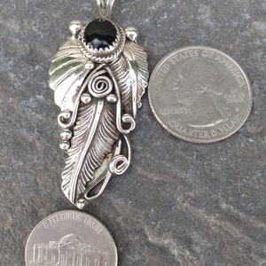 Puede incluir: Un colgante de plata con un dise&ntilde;o de pluma detallado y una piedra preciosa negra en la parte superior. El colgante se muestra junto a una moneda de 25 centavos de d&oacute;lar estadounidense para comparar el tama&ntilde;o.