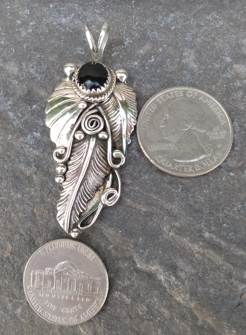 Puede incluir: Un colgante de plata con un dise&ntilde;o de pluma detallado y una piedra preciosa negra en la parte superior. El colgante se muestra junto a una moneda de 25 centavos de d&oacute;lar estadounidense para comparar el tama&ntilde;o.