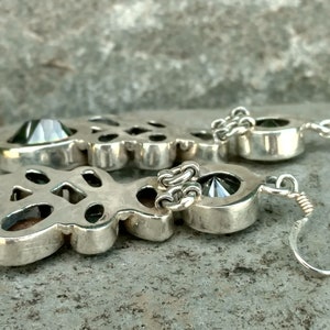 May include: A pair of silver earrings with a large, faceted, iridescent gemstone in the center of each earring. The earrings are connected to a silver chain and have a hook closure.
